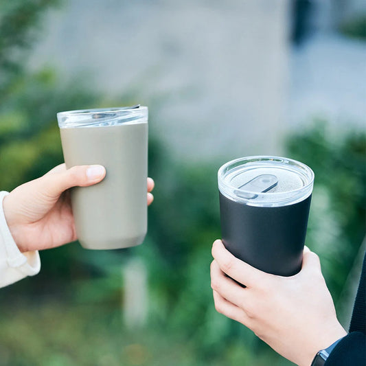 Two hands holding insulated travel mugs outdoors with a blurred green background