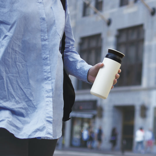 Person holding a white water bottle with a black lid on a city street.