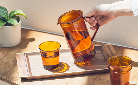 Hand pouring tea from a glass pitcher into two glasses on a wooden tray with a plant in the background.