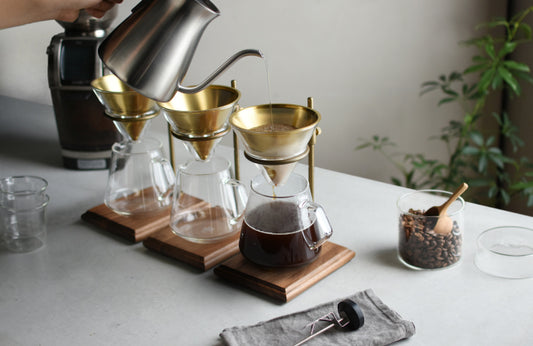 Person pouring coffee from a silver kettle into a glass carafe on a wooden coaster.