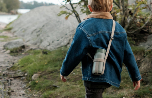 Child in a denim jacket with a backpack walking outdoors near a body of water.