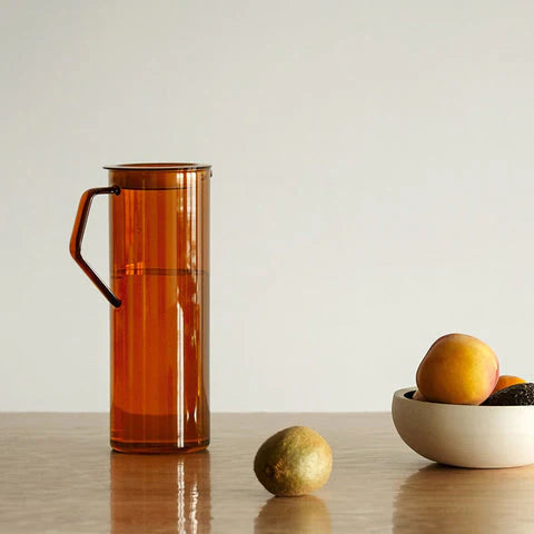Orange glass pitcher on a wooden surface with a bowl of fruit in the background