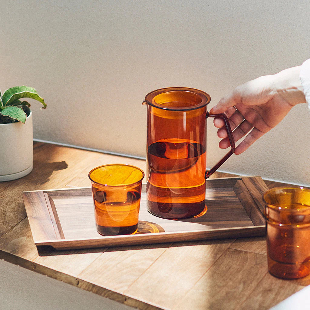 Person holding an amber glass pitcher on a wooden tray with two glasses.