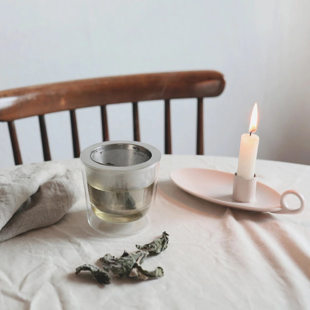 Clear glass of tea with a tea bag, dried tea leaves, and a lit candle on a wooden chair.