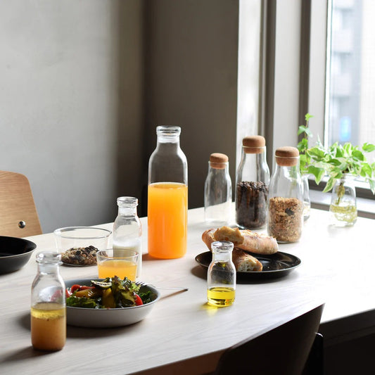 Table with various bottles, glasses, and food items near a window.