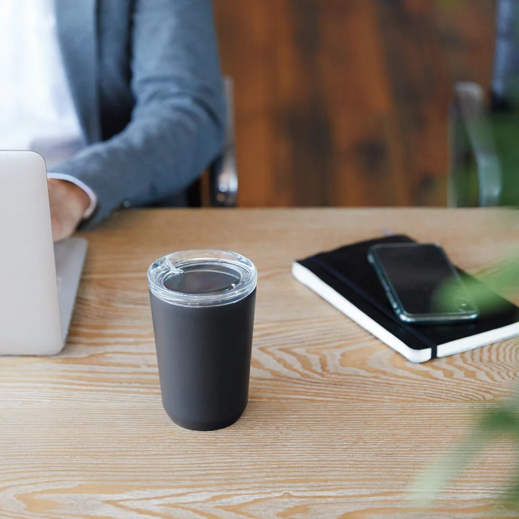 Black tumbler on a wooden desk with a person using a laptop in the background