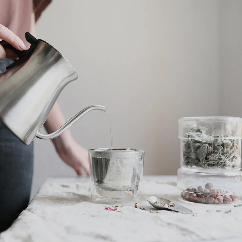 Person pouring tea from a silver kettle into a glass on a marble surface.
