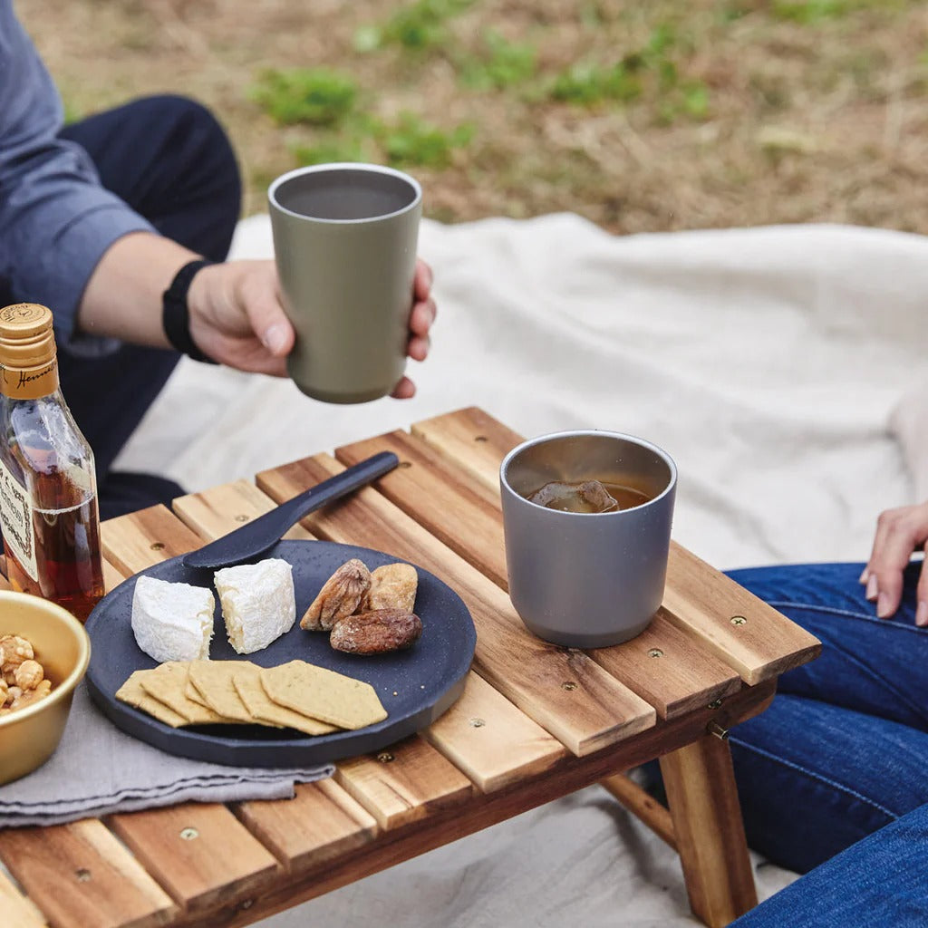 Person holding a mug with a small wooden table featuring food and drinks outdoors.