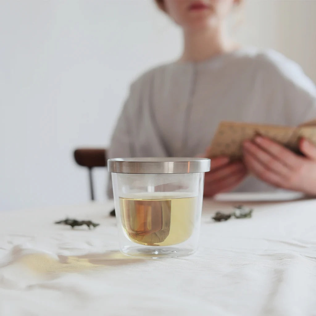 Clear glass cup with tea on a table, person reading a book in the background