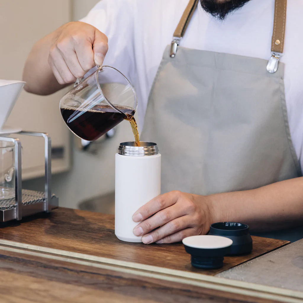 Person pouring coffee into a white thermos with a wooden counter and kitchen equipment in the background.
