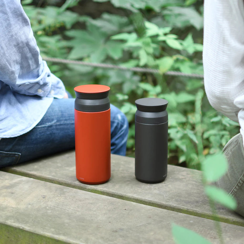 Two insulated travel mugs, one red and one black, on a wooden surface with a natural background.